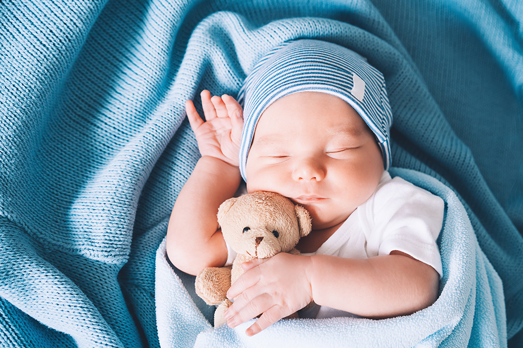 A baby with shoulder dystocia is wrapped in a blue blanket and hat, sleeping while holding a teddy bear.